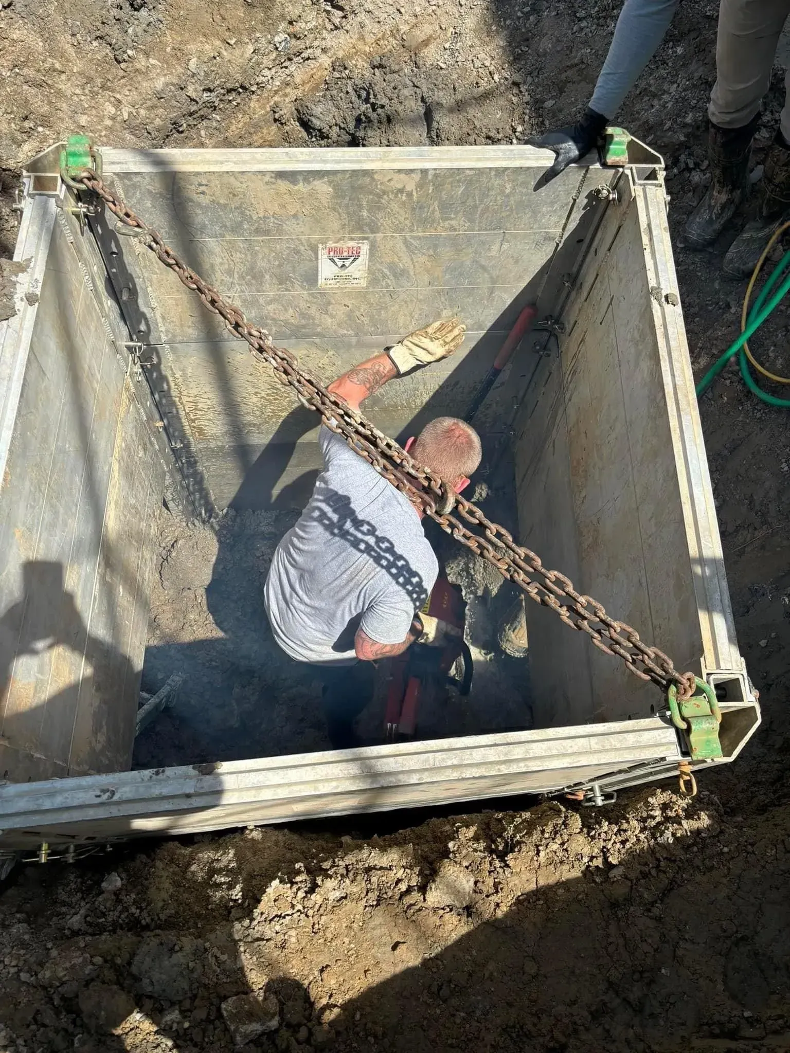 Man in trench box in a dirt excavation, held by chains, one person assisting from above.