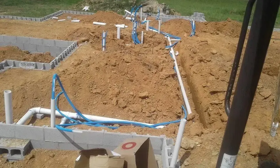 Construction site with exposed plumbing in dirt, near a cinder block foundation.