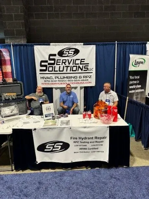 Three men at a trade show booth for Service Solutions LLC. They are behind a table with a banner about HVAC, plumbing, and fire hydrant repair.