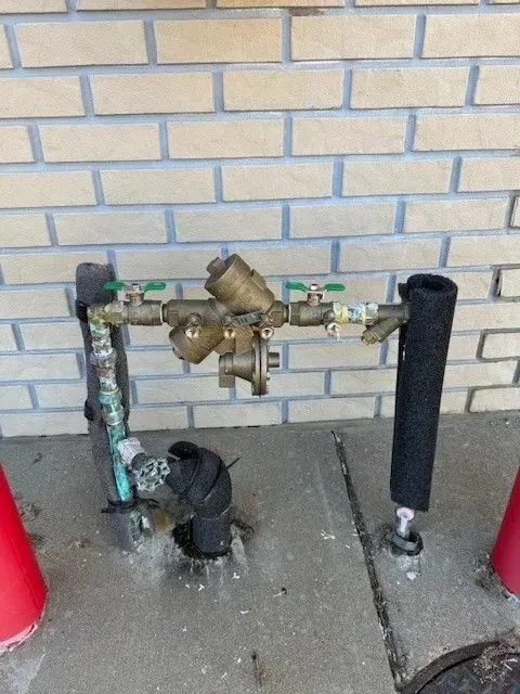 Water backflow preventer with valves, pipes, and insulation against a brick wall.