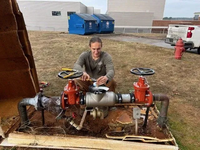 Man working on water pipes outdoors near a building, with tools, red valves, and a brown wooden enclosure.