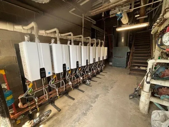 Row of white water heaters in a basement; pipes and stairs visible.