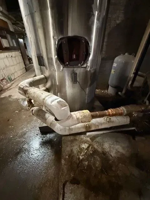 A large, stainless steel tank with a pipe, possible water damage, and a water heater in a basement setting.