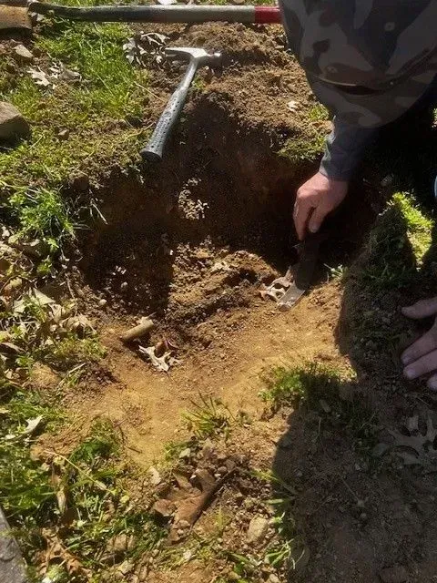 Person digging in a hole in the ground, a hammer lies nearby.