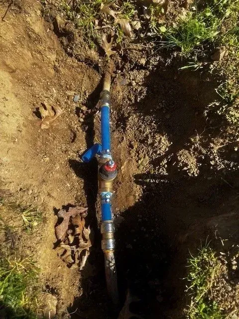Water valve and pipes in a hole in the ground, blue and metal components, surrounded by dirt and grass.