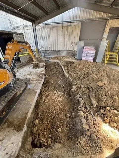 An excavator digs a trench inside a building. Soil piles up next to the trench.