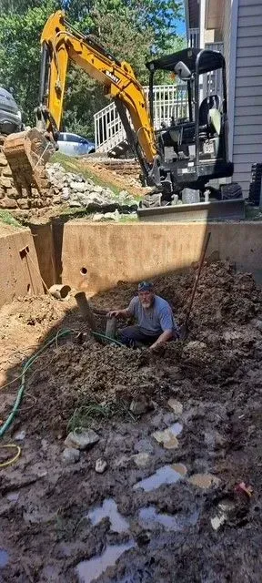A man is working in a muddy trench with an excavator in the background.