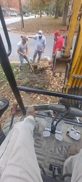 Three men working on a dig site; one operates an excavator, others assist.  Autumn leaves on the ground and trees.