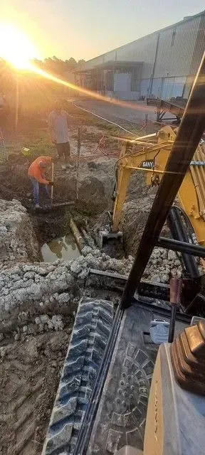 Construction site. Workers excavating a trench near a warehouse with an excavator and shovels. Sunset in the background.