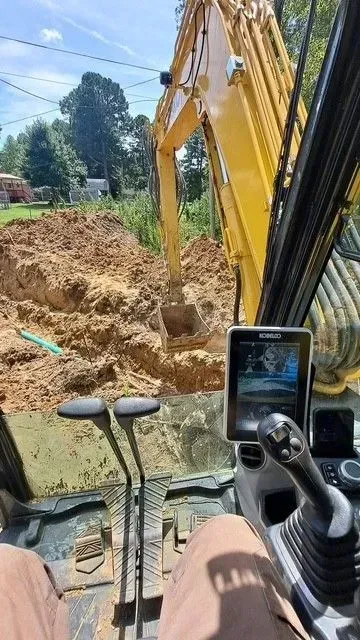 Inside excavator cab, operating arm digging into dirt. Yellow machine, brown soil, sunny day.