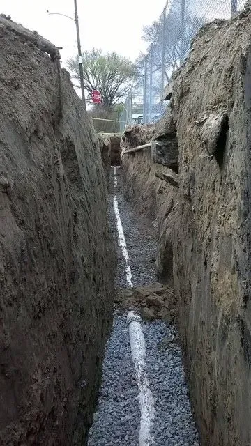 Trench dug for laying a white pipe, surrounded by dirt walls, in an outdoor setting.