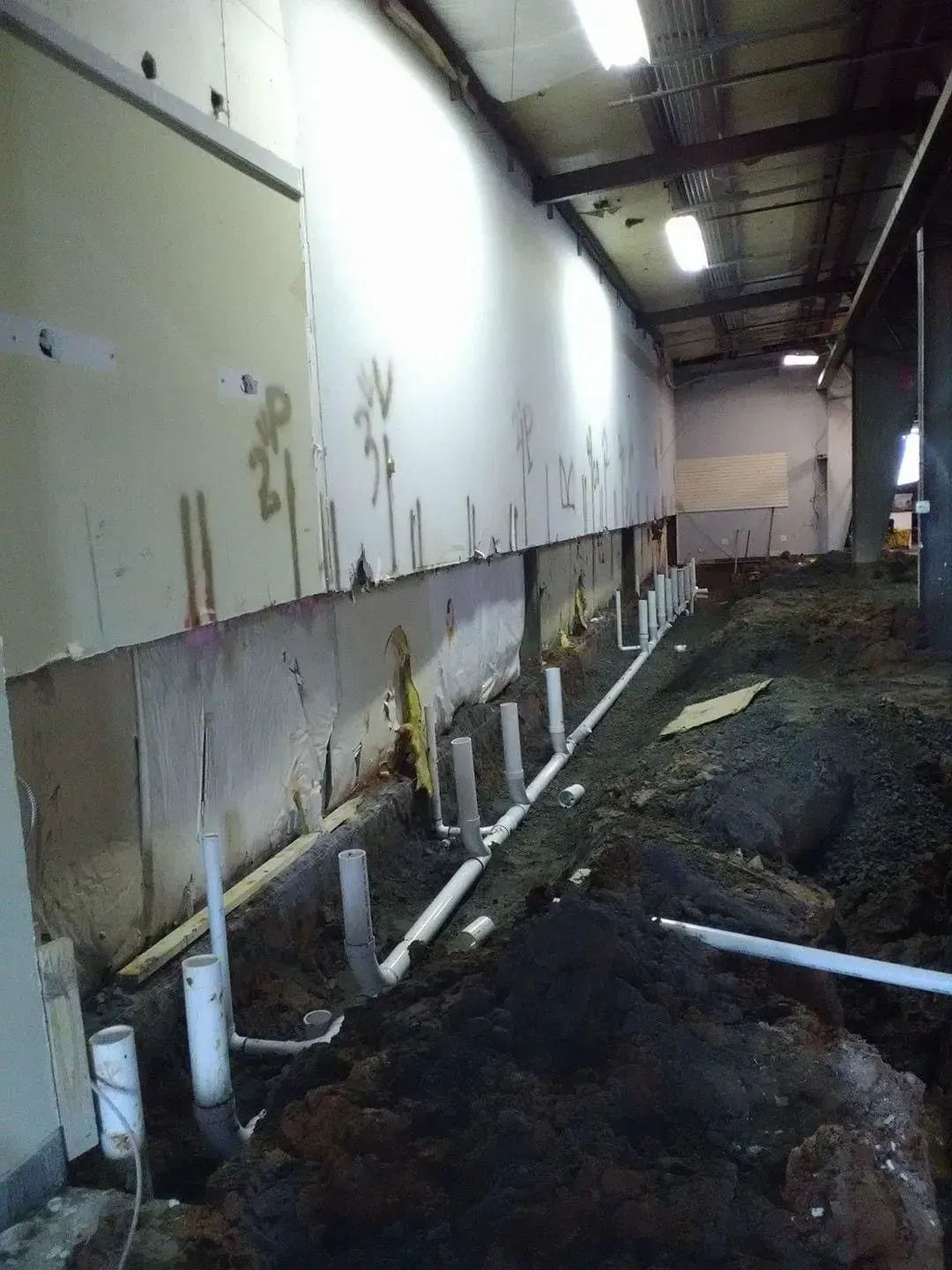 Construction site: Exposed pipes and dirt along a wall. White pipes, brown dirt, and a mostly white wall.