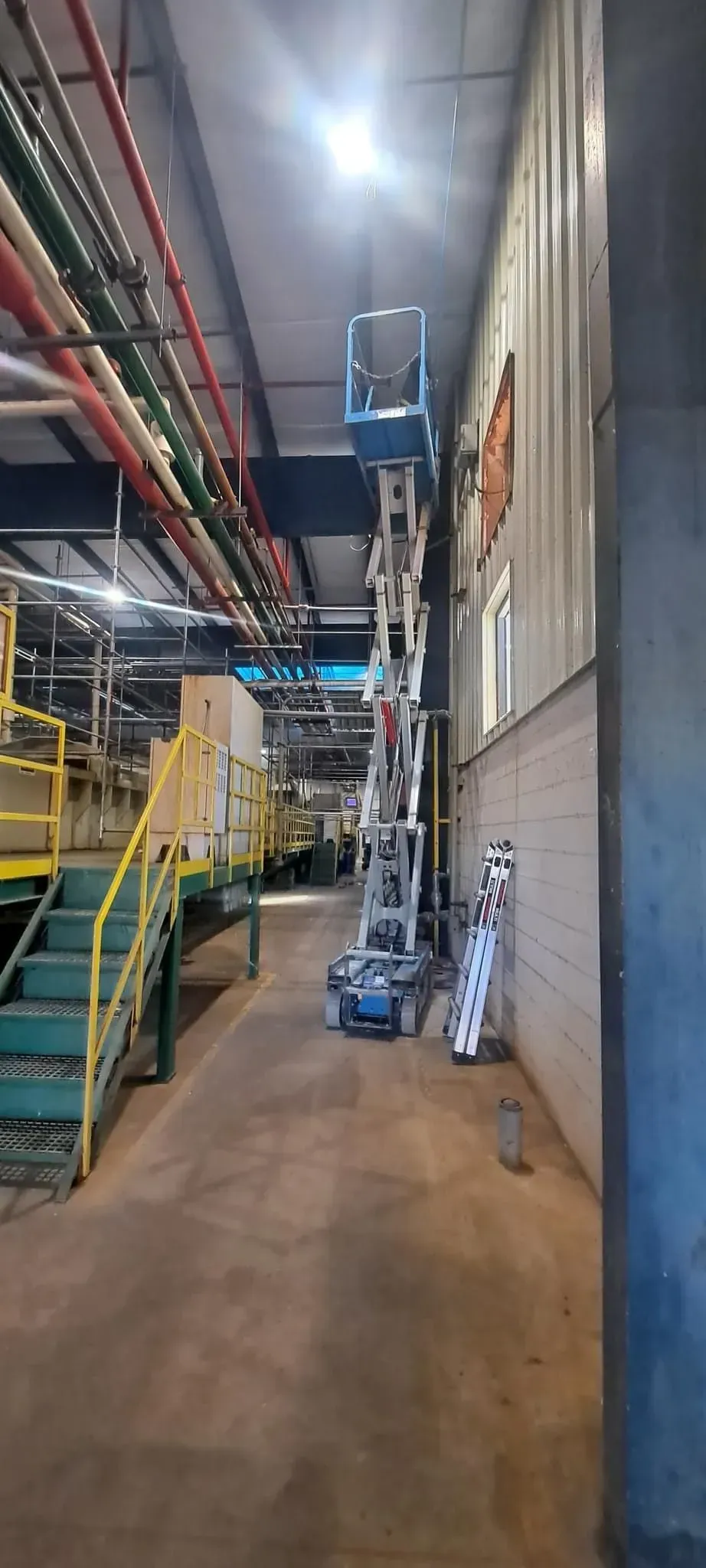 A lift is raised next to a factory wall. Staircase and pipes are on the left.