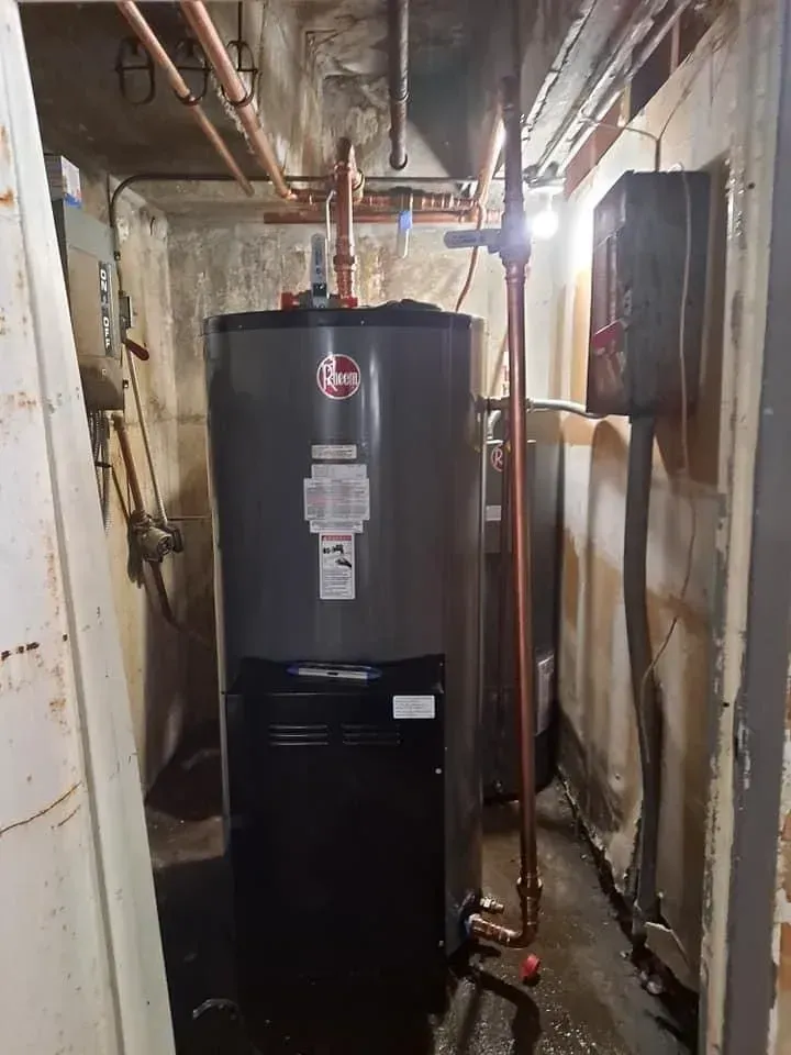 A tall, dark water heater in a cramped, damp basement. Copper pipes and a control box are visible.