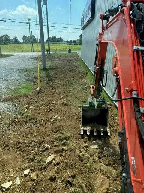 Orange excavator digging next to a building. Ground is dirt and rocks. Blue sky in background.