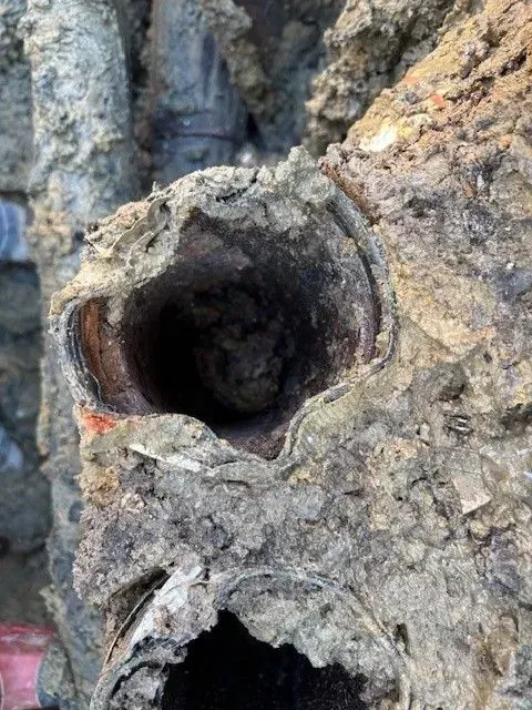 Severed, corroded pipe in soil, revealing dark interior, surrounded by mud and other pipes.