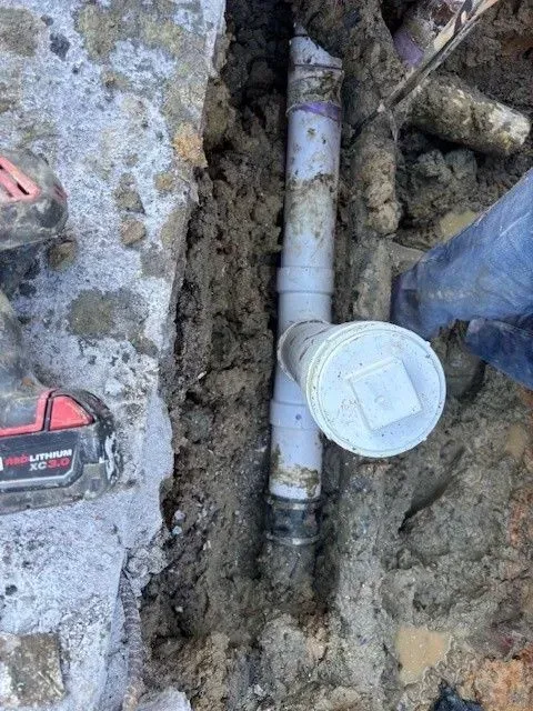 Plumbing pipes in a trench; a person is working on them with a drill.