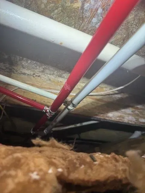 Pipes in a crawl space. Red and white PEX pipes connect to other pipes. Insulation is visible below.