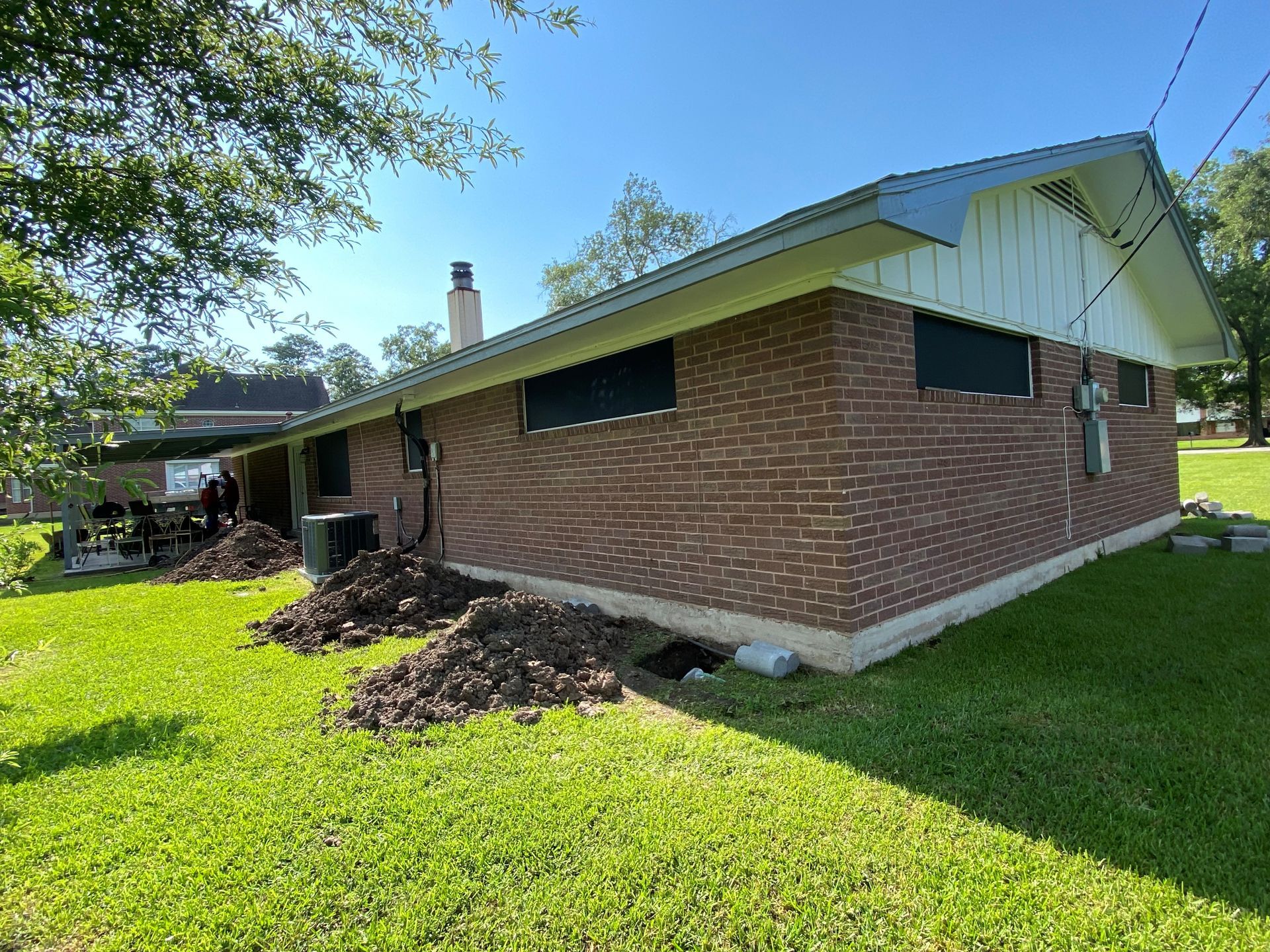 Brick house with dirt piles on the lawn. Green grass, blue sky, and a few trees are in the background.