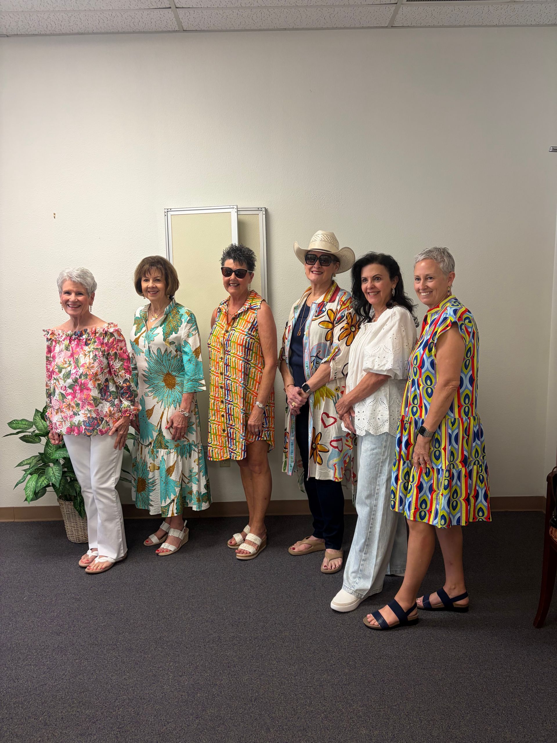 Six people stand in a line against a plain white wall, wearing colorful patterned tops and dresses.