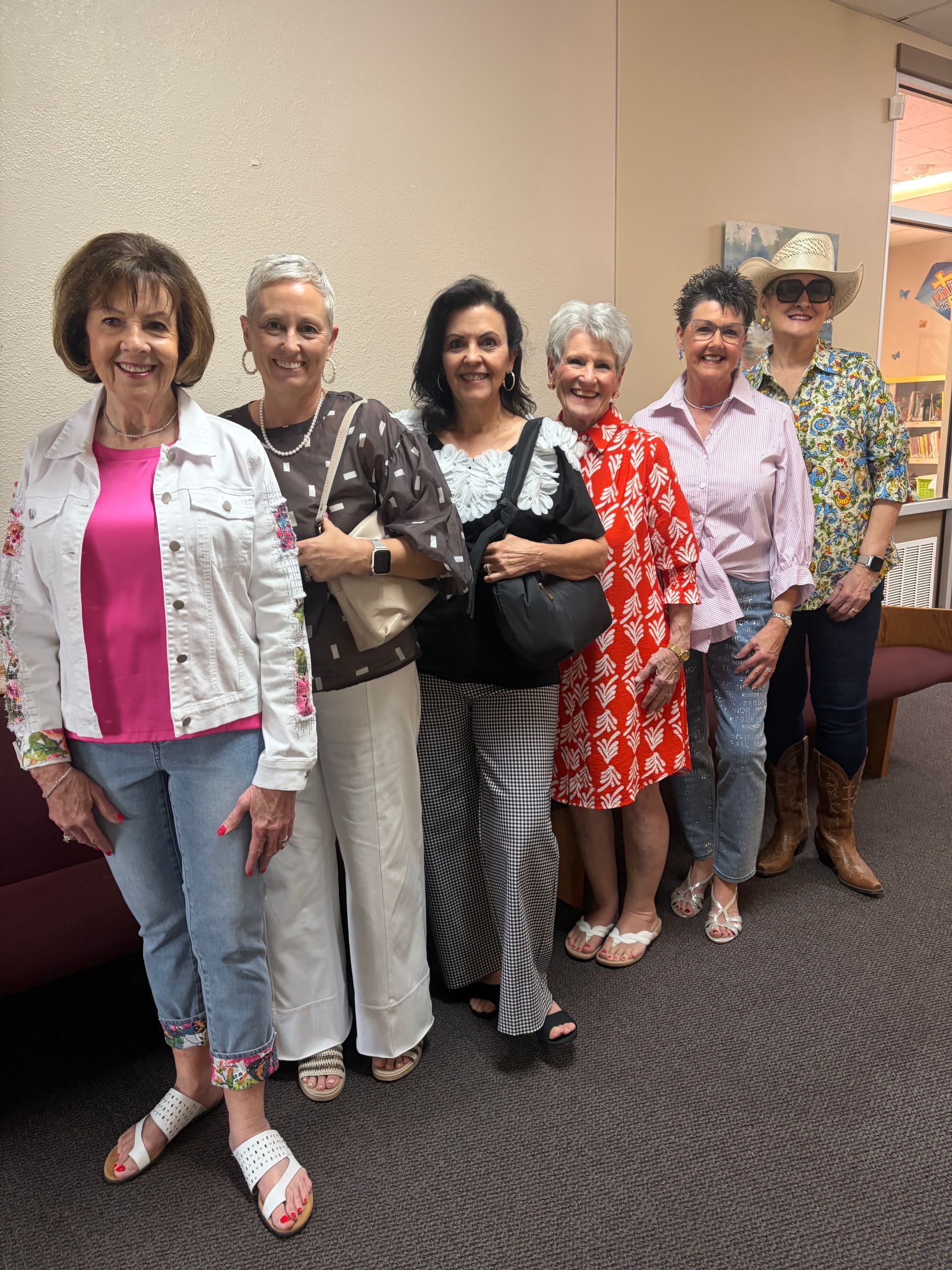 Six people stand in a line against a wall, smiling, dressed in a variety of colorful casual and western-themed clothing.