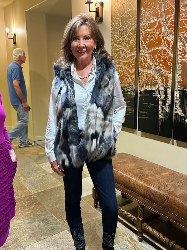 A woman wearing a fur vest and jeans is standing in a hallway.