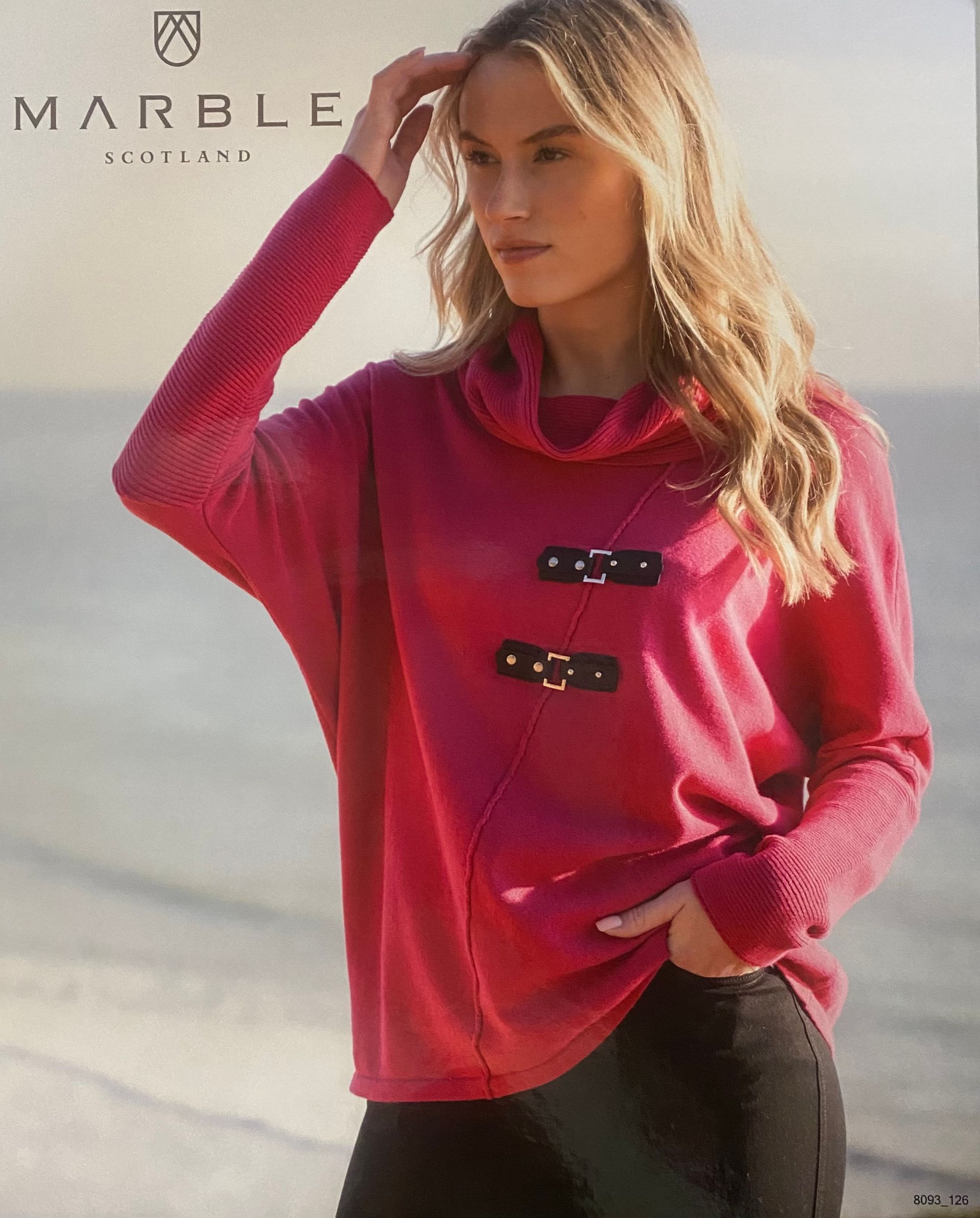 Woman in pink sweater with toggle closures, standing by the sea.