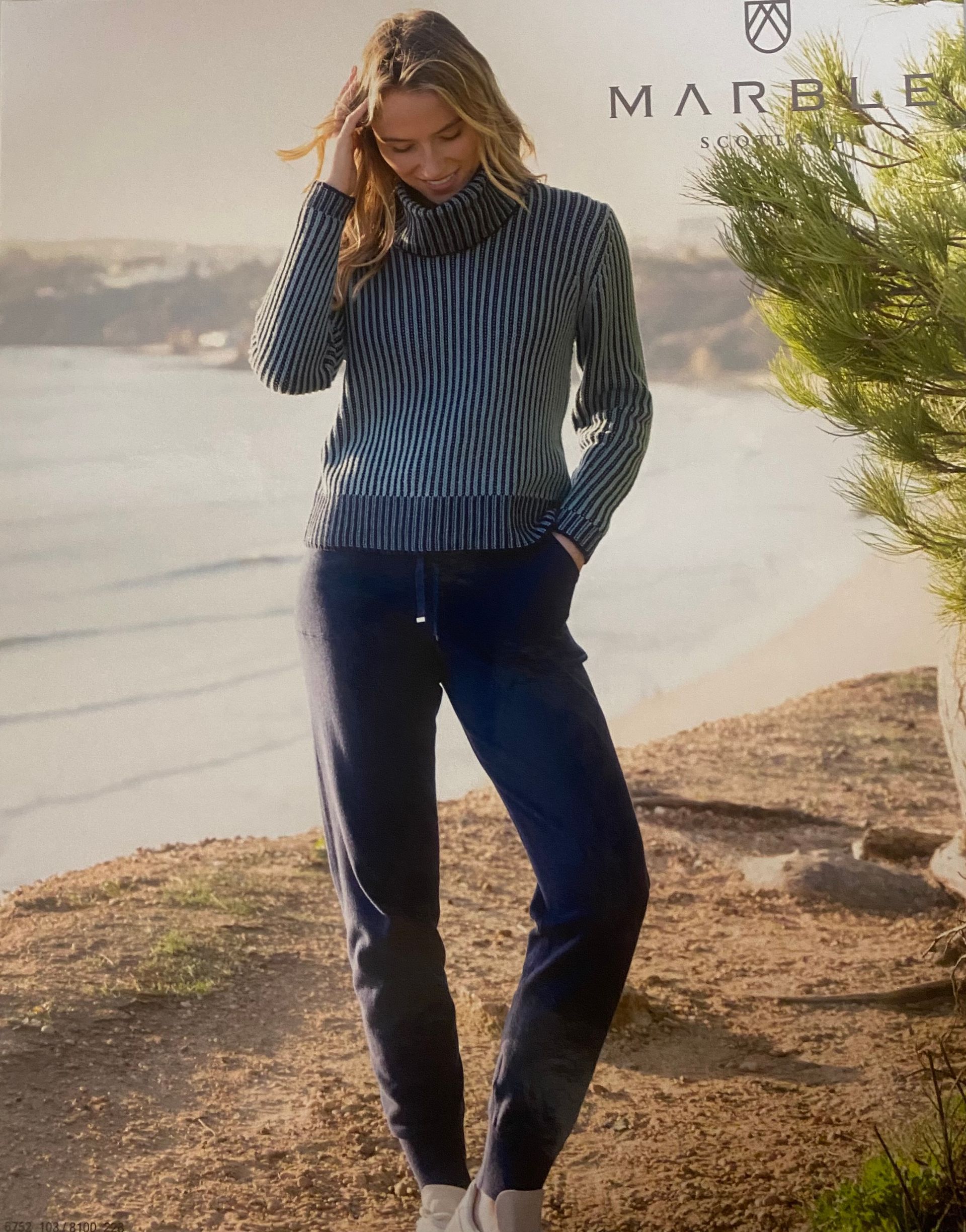 Woman in striped sweater and navy pants stands outdoors, beach in background.