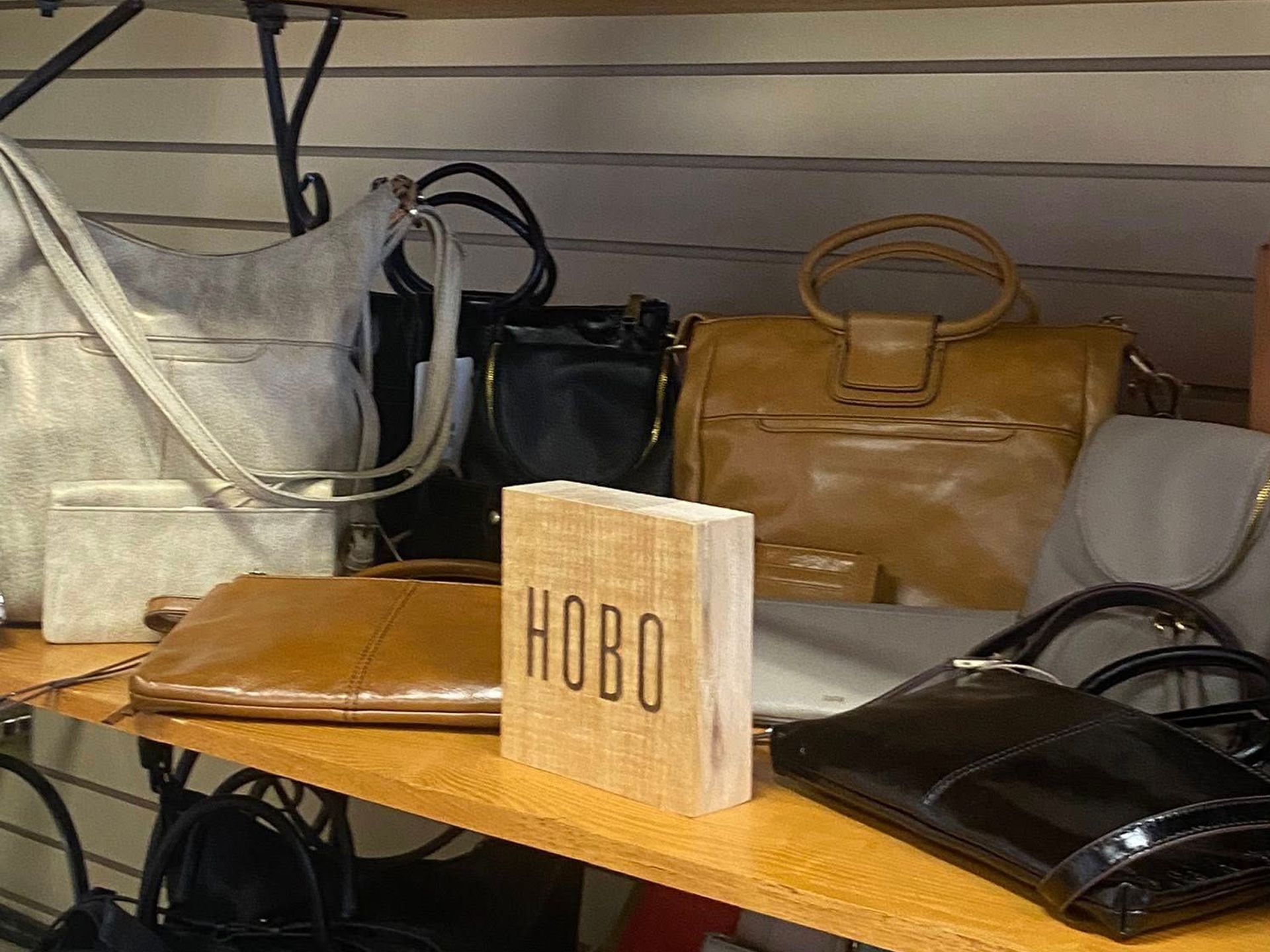Various leather HOBO handbags are on a shelf, including purses and shoulder bags in colors like brown, black, and gray.