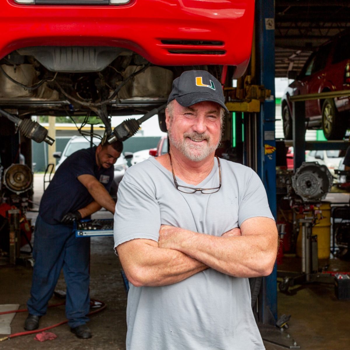 Mechanic workshop with a red car raised on a lift; man in gray shirt stands arms crossed in front.
