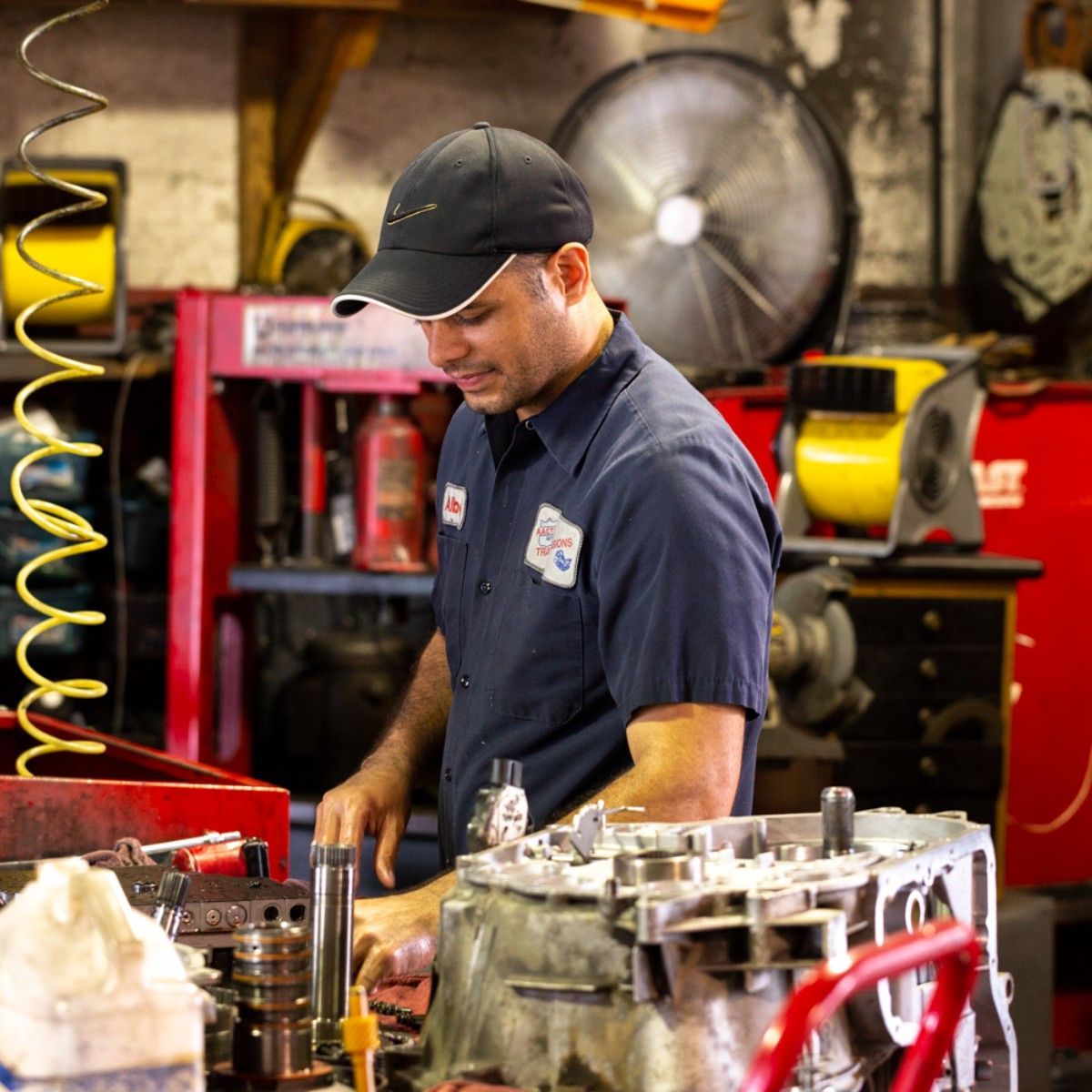 Mechanic in a dark blue shirt inspecting engine parts in a workshop, with tools and equipment around him