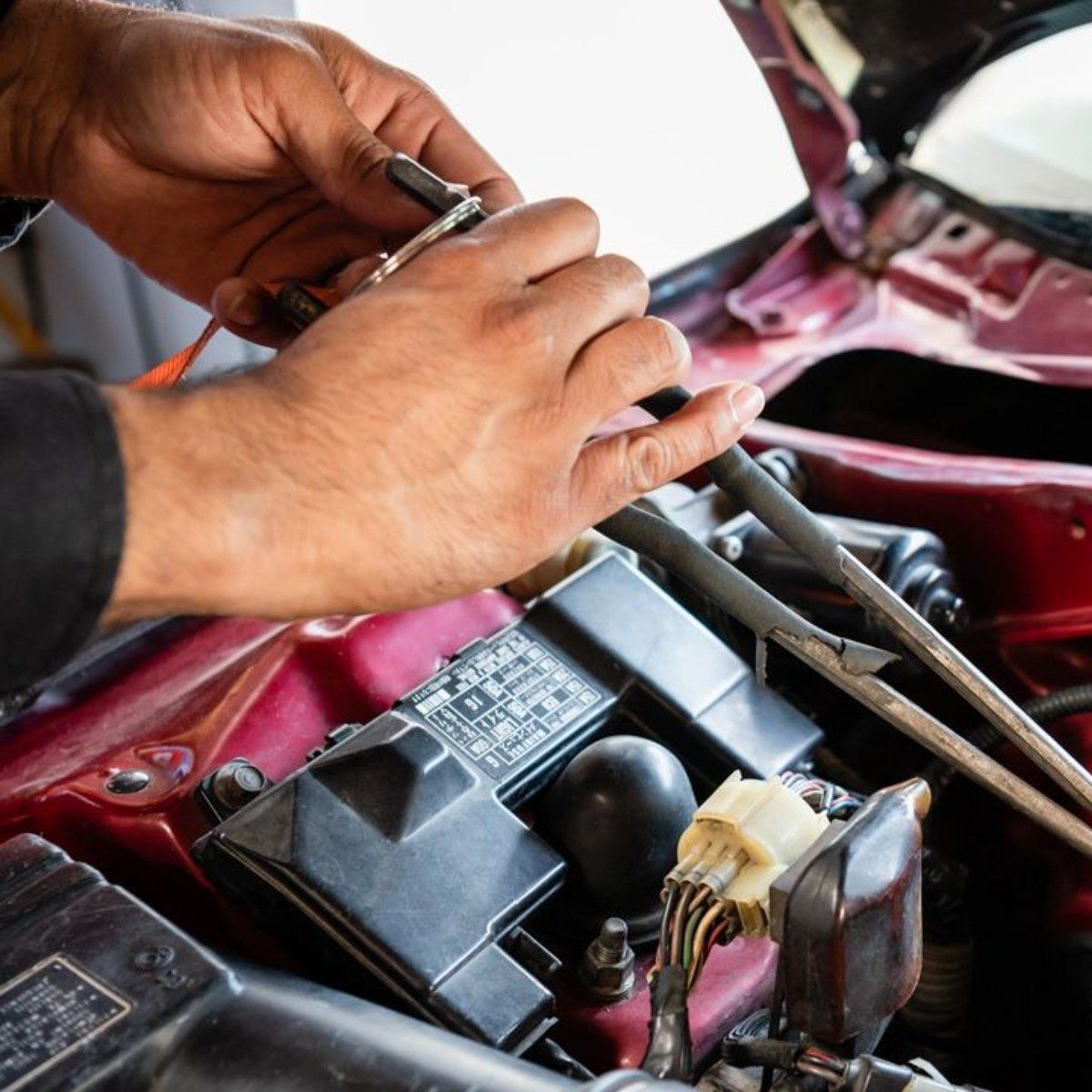 Hands working on car engine wiring in a maroon vehicle’s open hood compartment