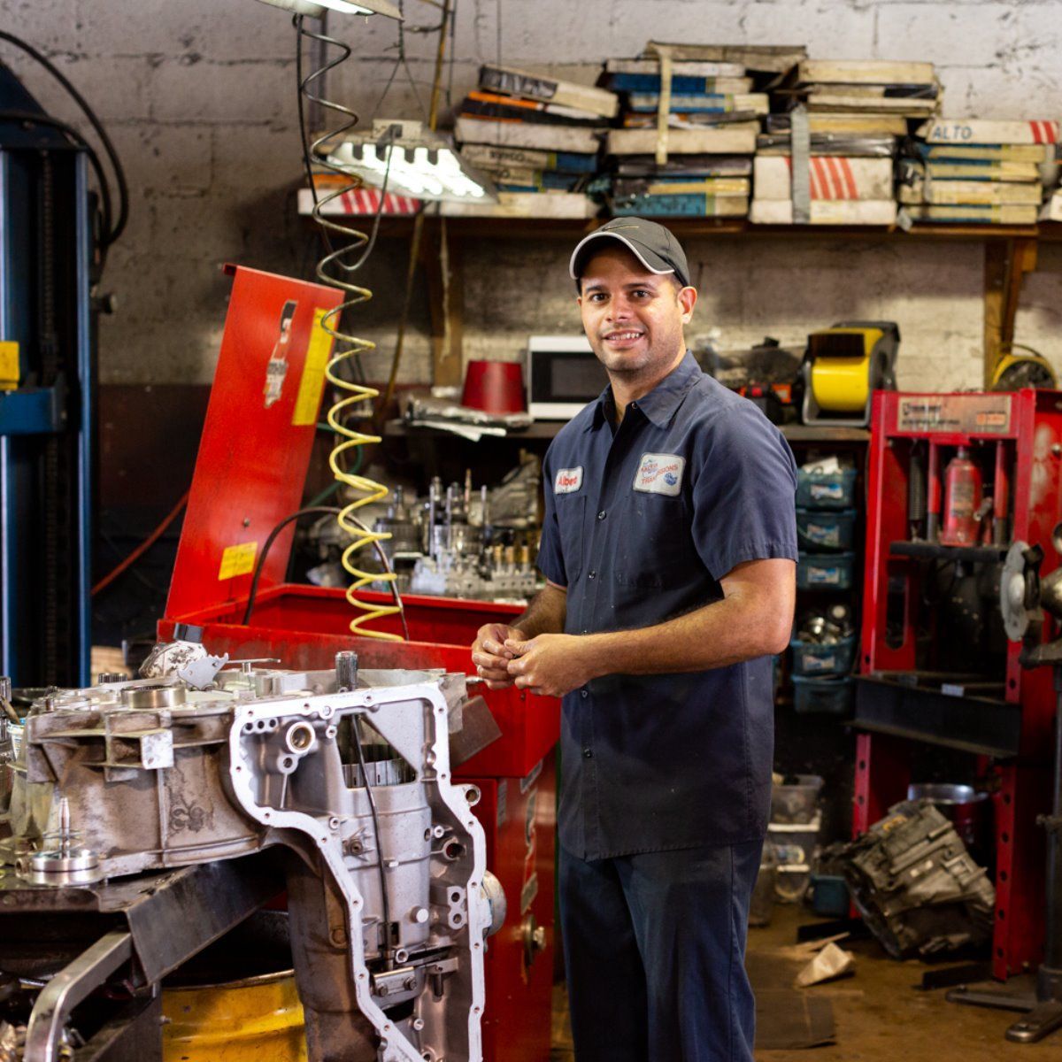Mechanic in a red-and-black auto repair shop working beside an engine block on a red lift