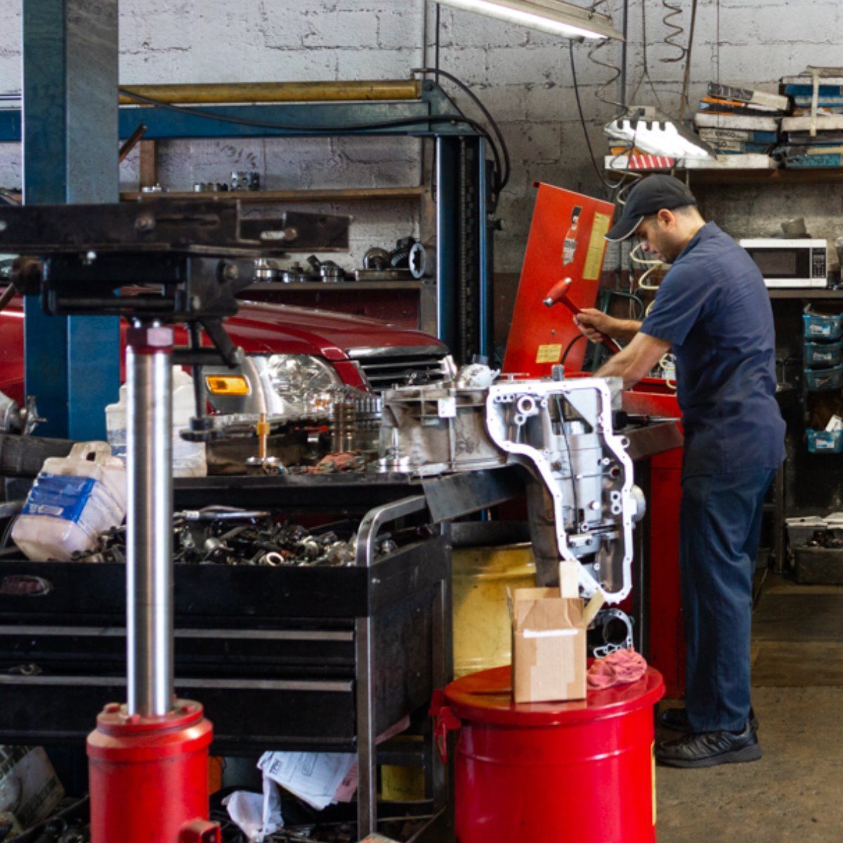 Mechanic working beside a red car in an auto repair shop with tools and equipment.