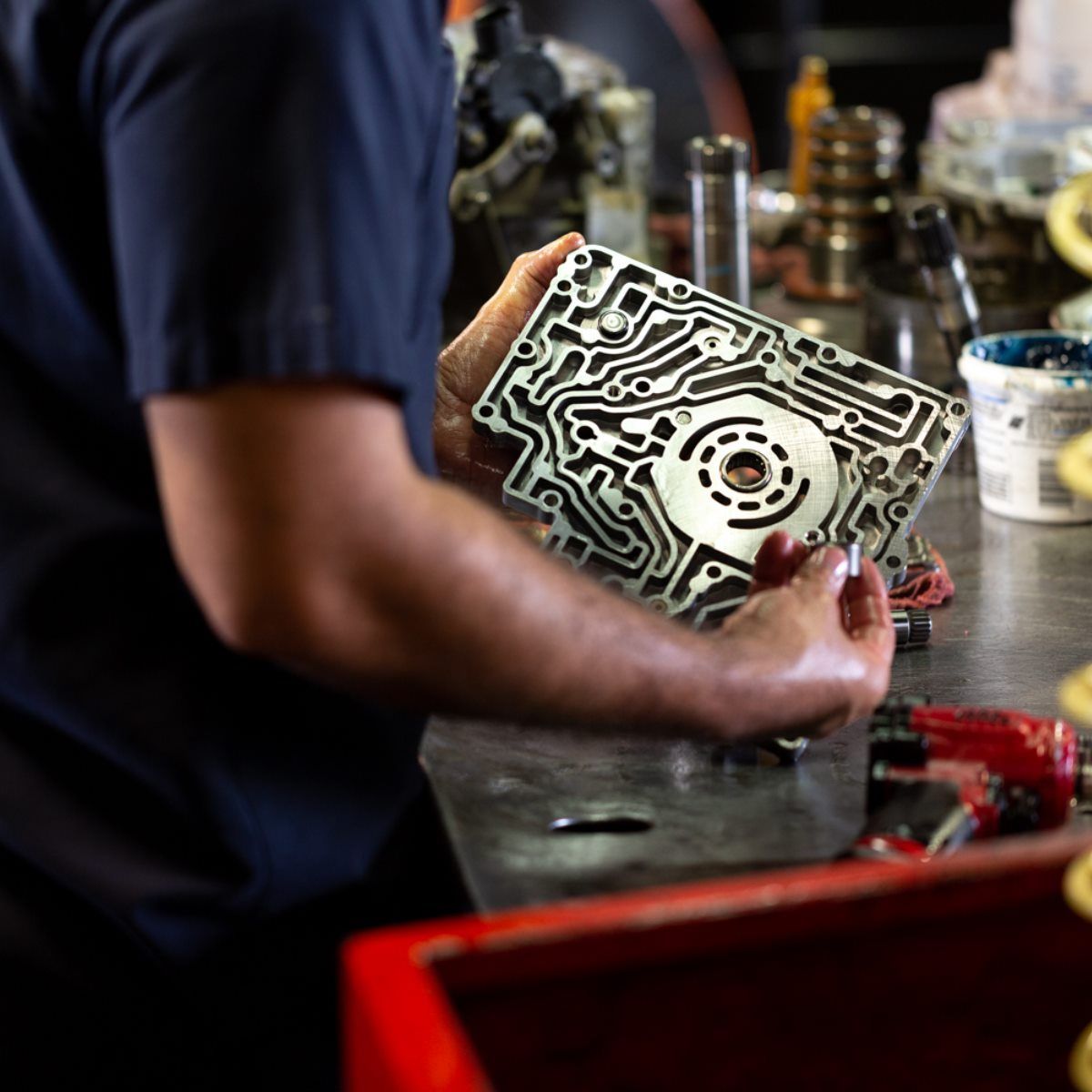 Mechanic inspecting a metal engine part at a workshop bench, with tools and containers in the background