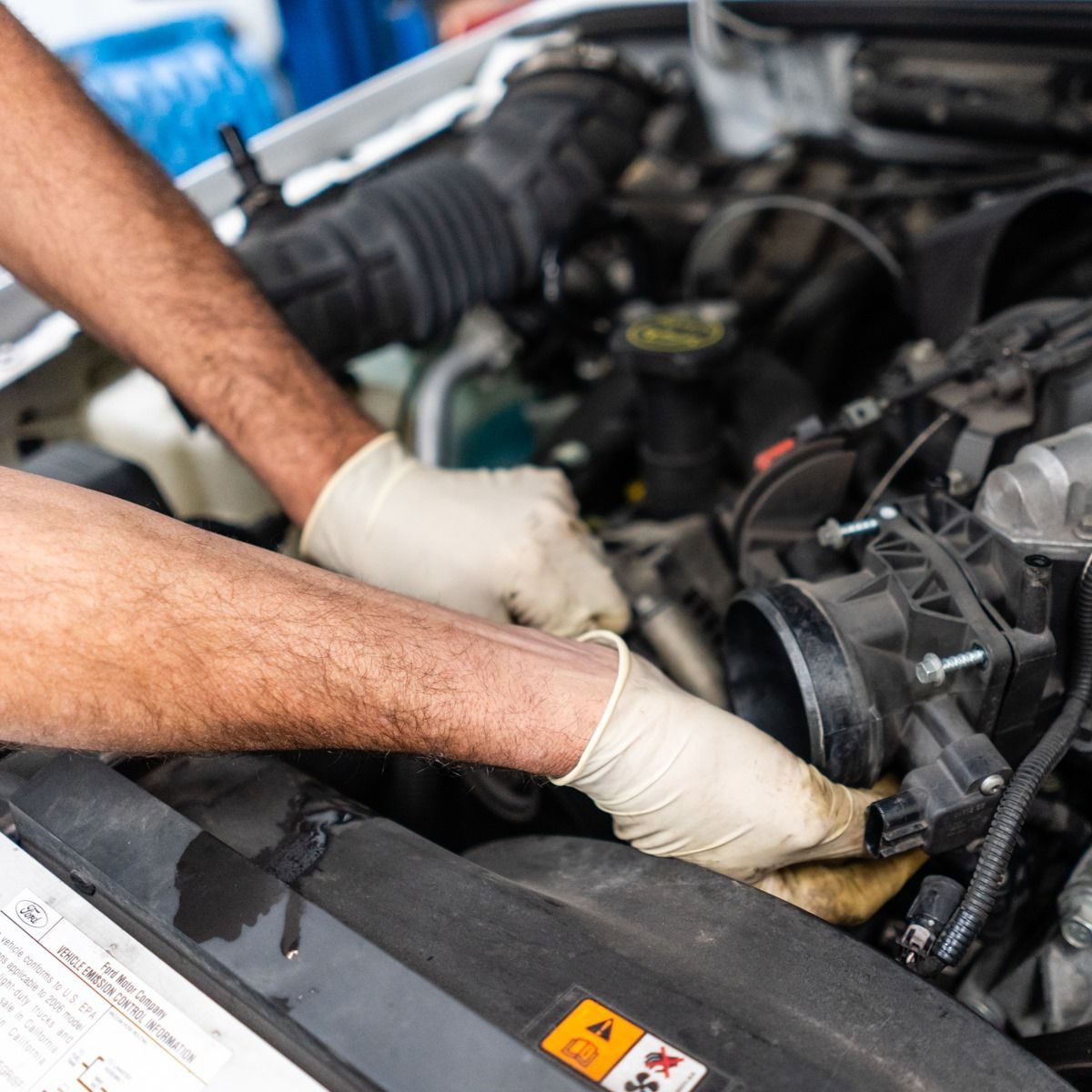 Mechanic wearing gloves works under a car hood, inspecting the engine bay.