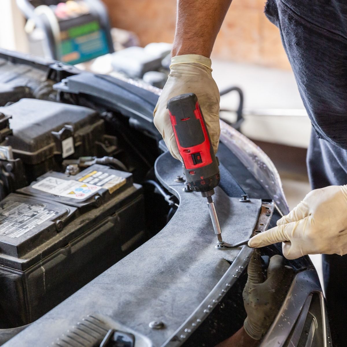 Mechanic in gloves using a red power drill to work on a car’s front panel under the hood