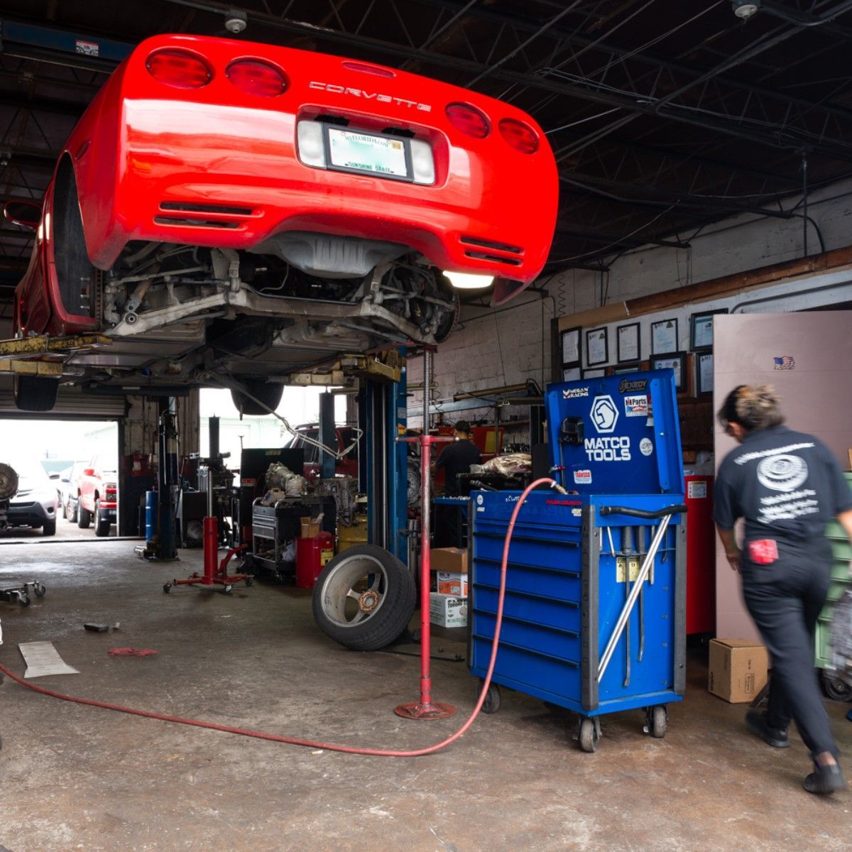 Red sports car lifted in an auto repair shop, with tools and a mechanic nearby.