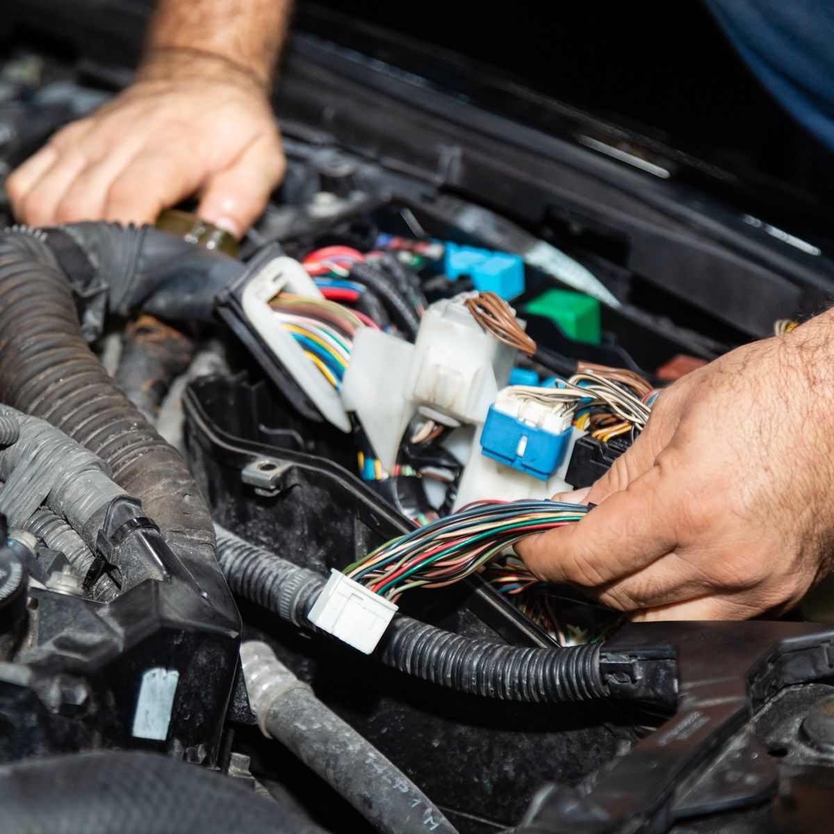Hands repairing exposed car wiring under the hood with connectors and cables visible.