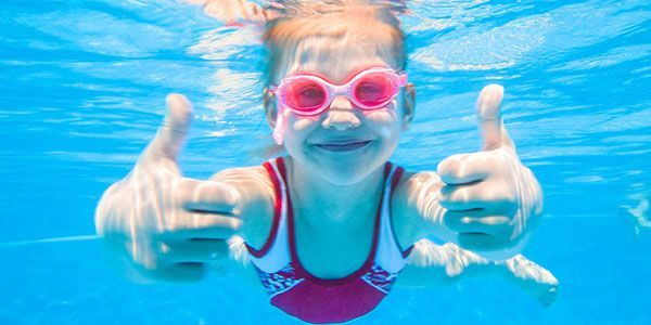 A little girl is swimming underwater in a pool and giving a thumbs up.