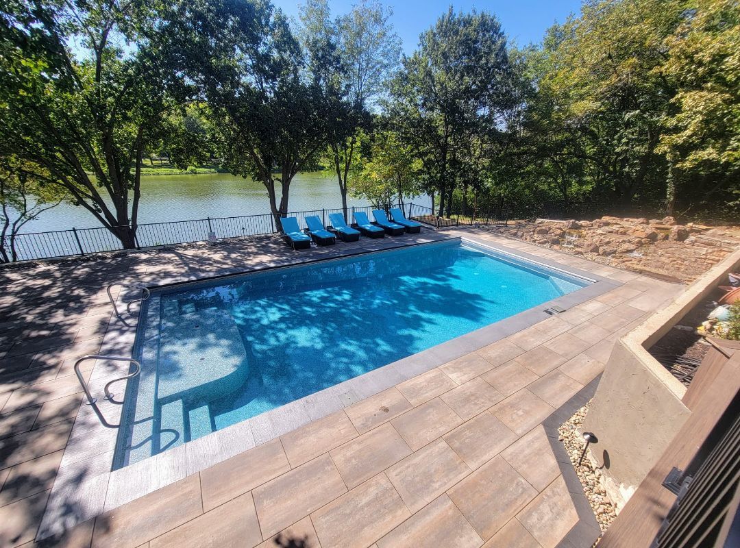 A large swimming pool surrounded by trees and chairs next to a river.