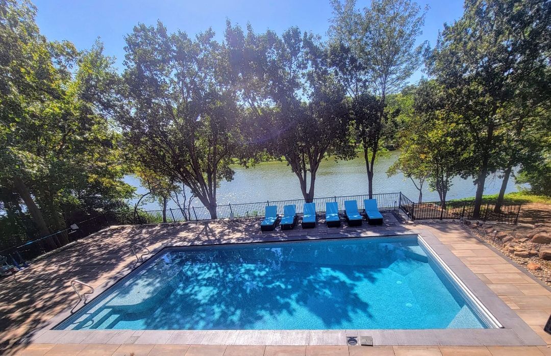 A large swimming pool surrounded by trees and chairs next to a lake.