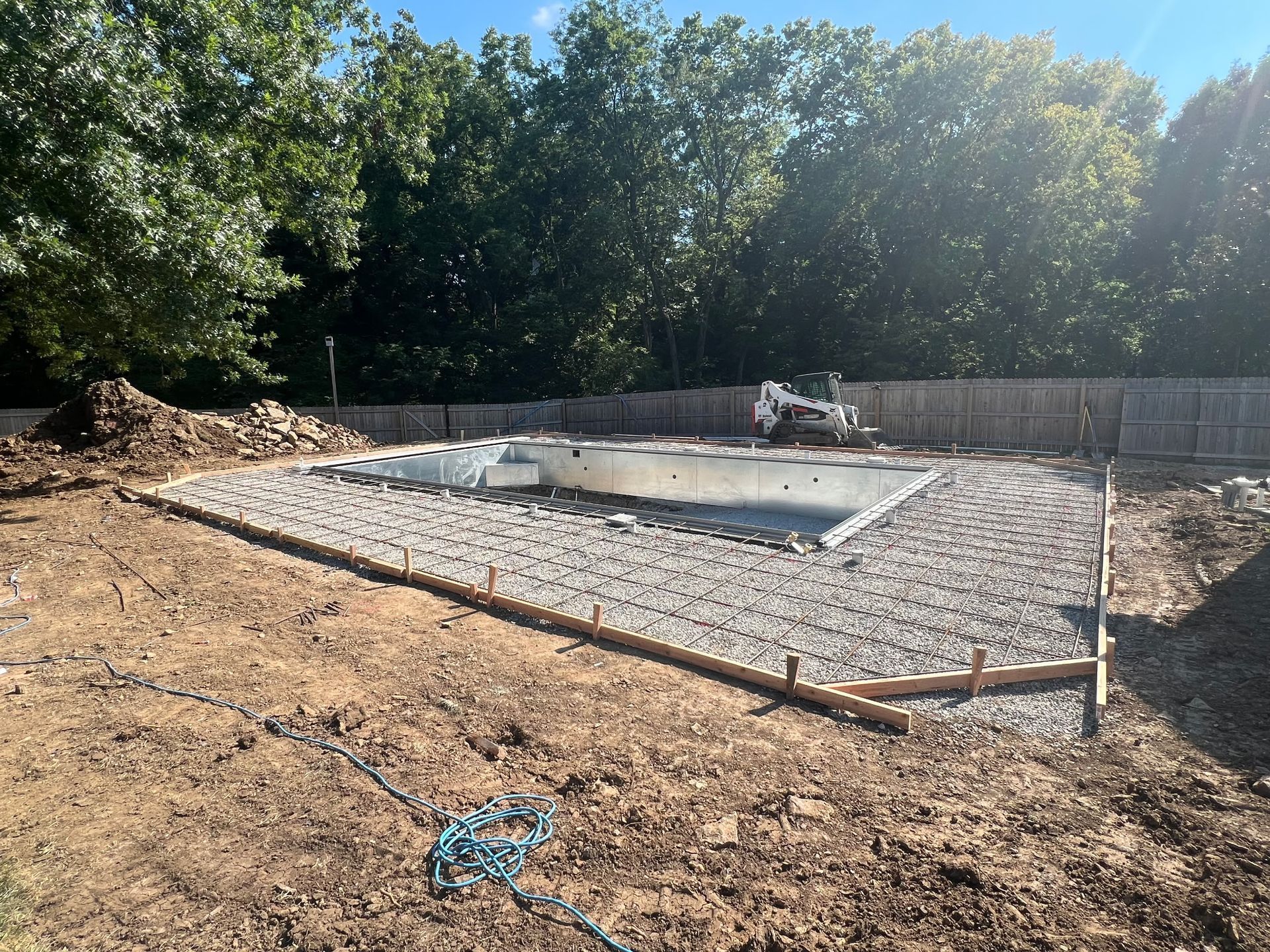 pool construction site with gravel base and excavator, surrounded by dirt and trees