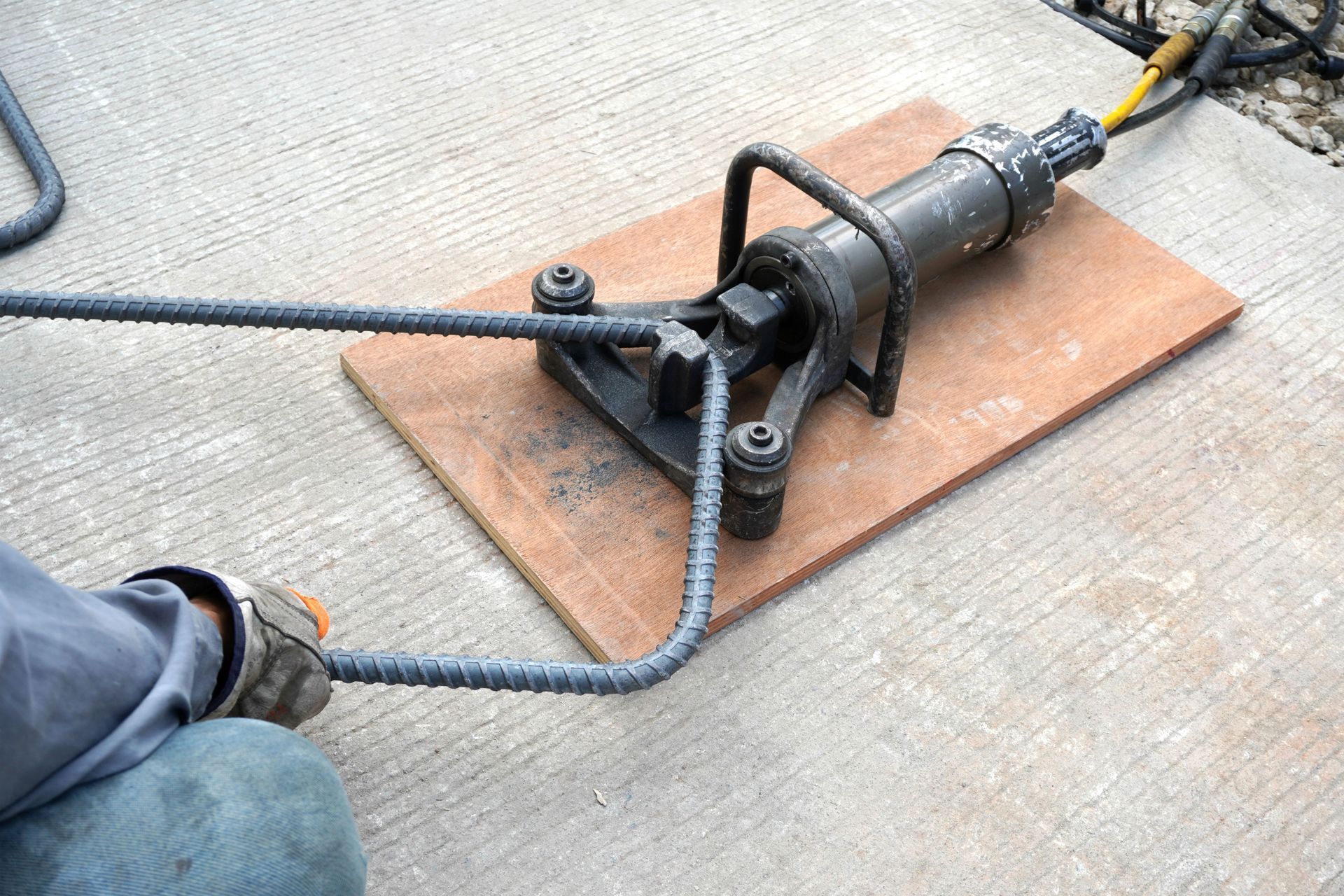 Person bending rebar with a hydraulic bender on a plywood base, outdoors.