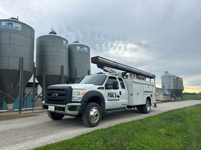 White utility truck driving past grain silos on a rural farm road.
