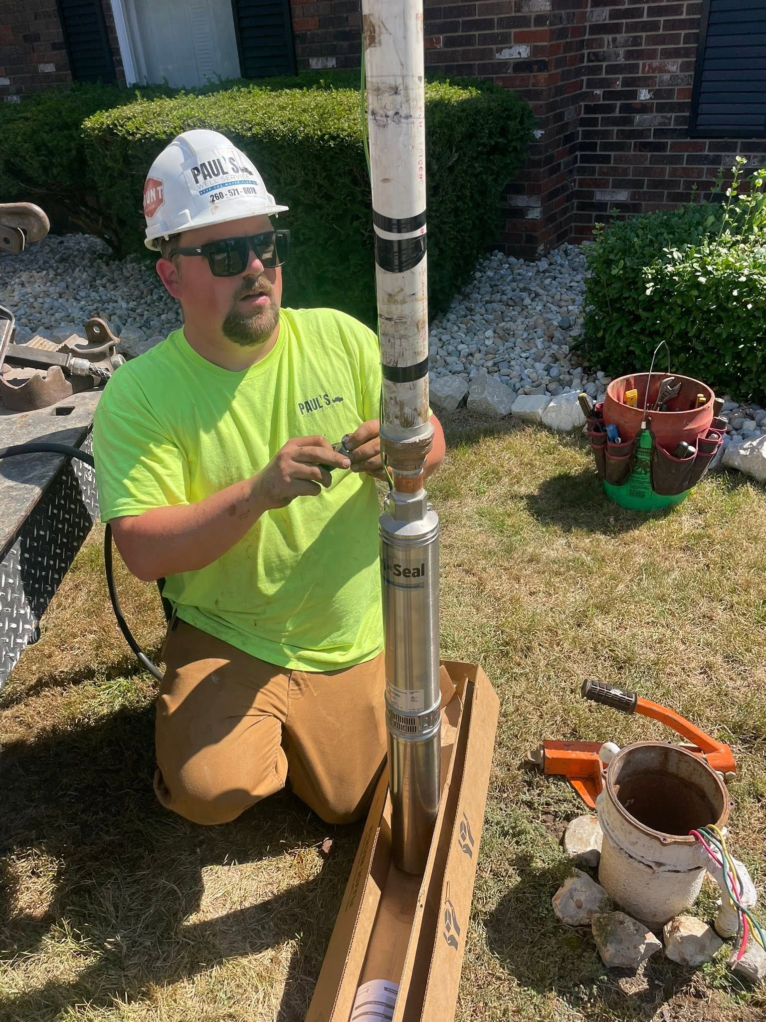 Worker kneeling on lawn, installing a metal pipe assembly beside a brick house and garden tools.