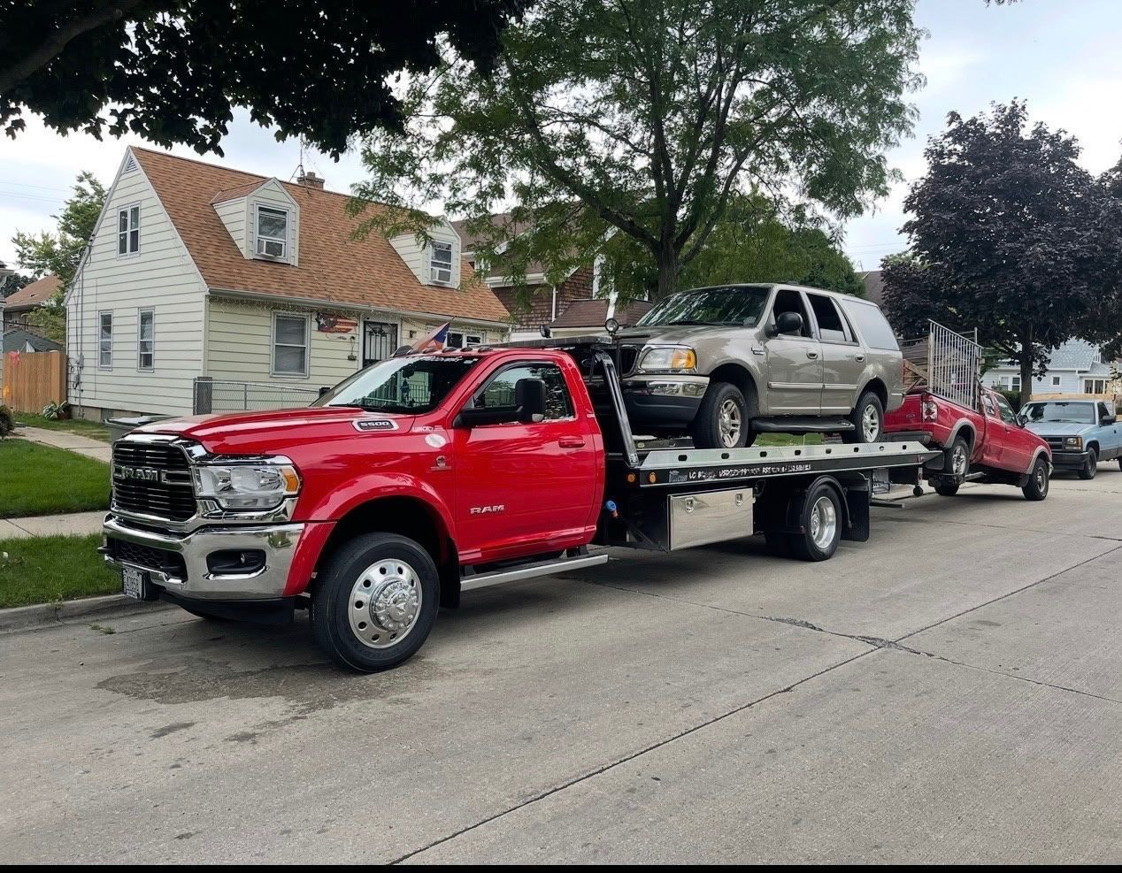 Red tow truck carrying two cars on a city street in front of houses.