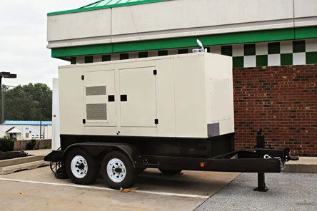 A generator on a trailer is parked in front of a brick building.