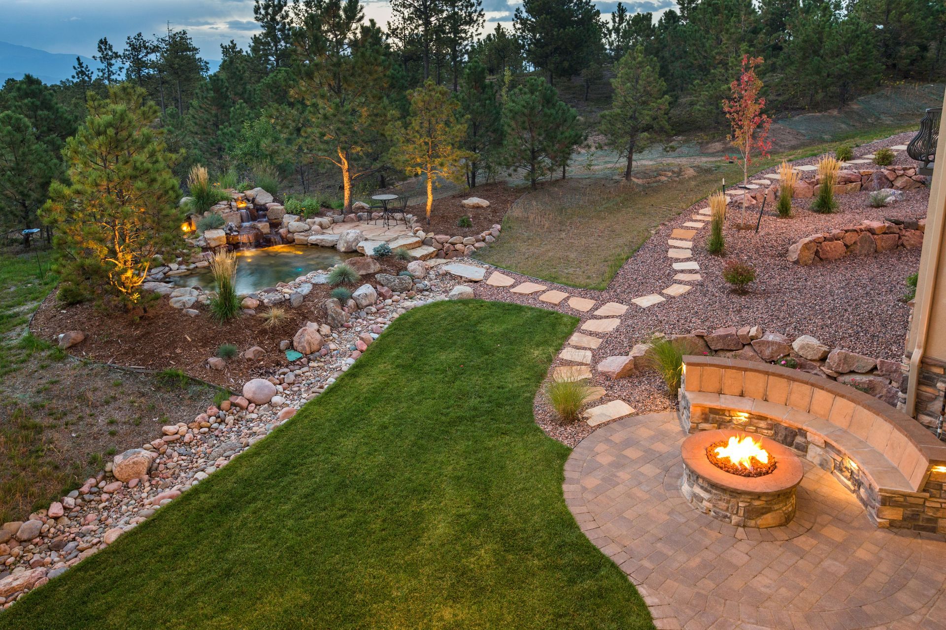 Backyard patio with fire pit, stone path, lawn, and small waterfall garden at dusk
