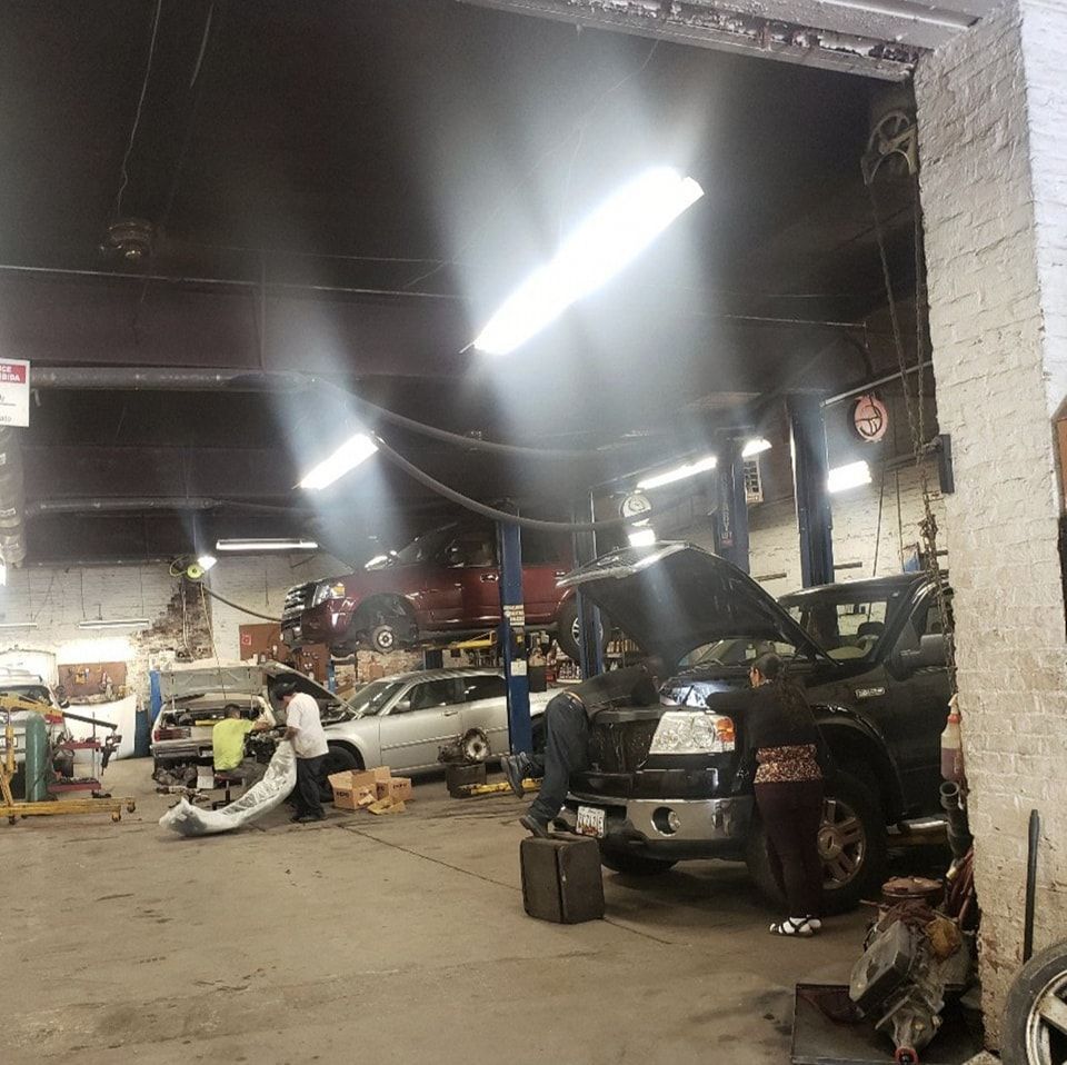 Mechanics work on vehicles inside a busy automotive repair shop with exposed ceiling beams and bright shop lights.